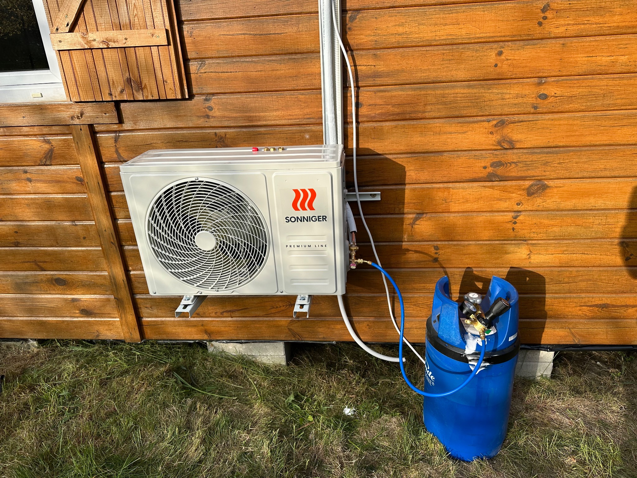 Montaż klimatyzacji zewnętrznej Sonniger na drewnianej elewacji domu. Widoczna butla z gazem i przewody. Instalacja na trawniku, w słoneczny dzień.