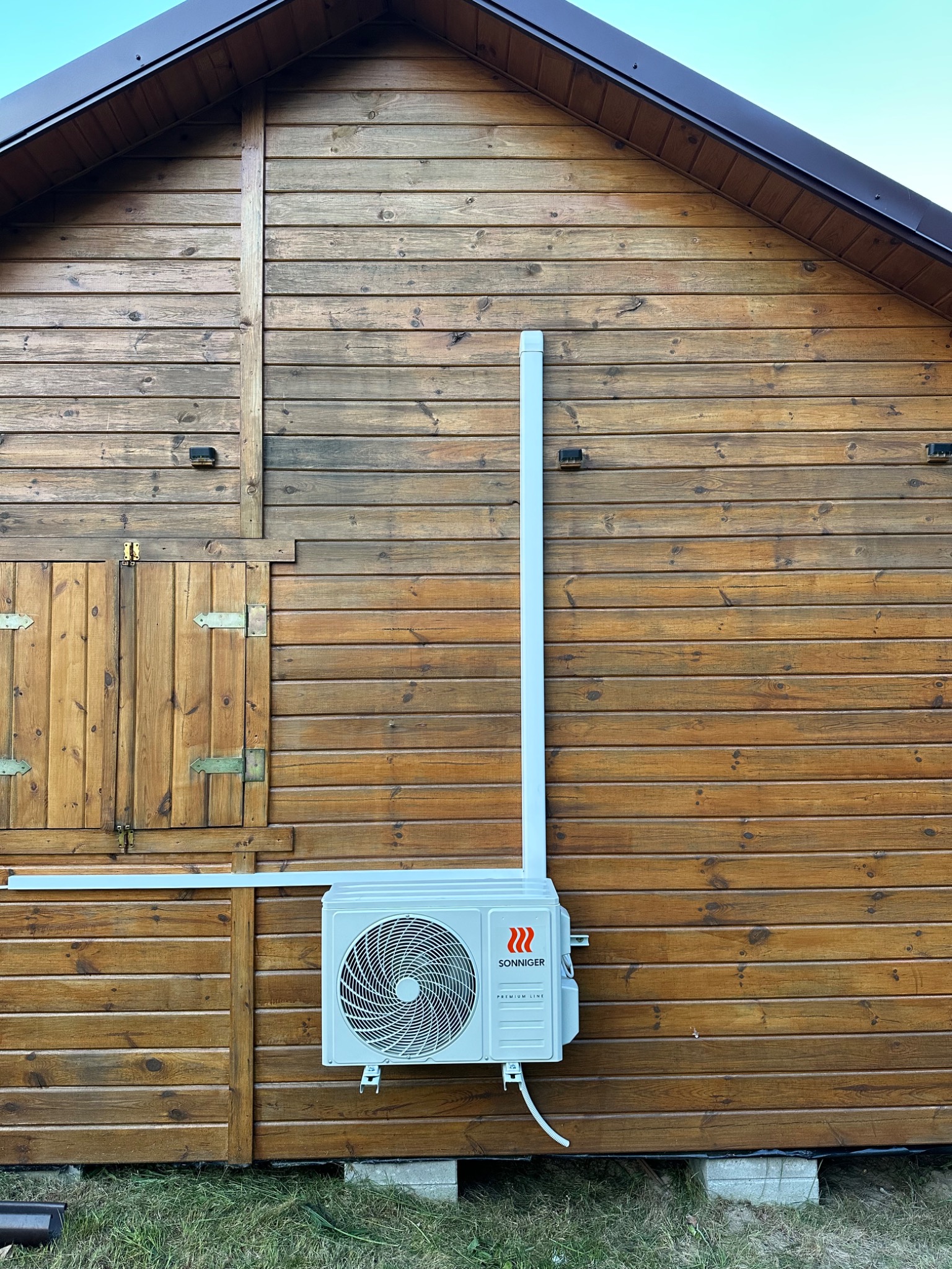 Biała klimatyzacja zewnętrzna marki Sonniger zamontowana na drewnianej ścianie domu, z widoczną instalacją odprowadzającą. Ujęcie z dołu.