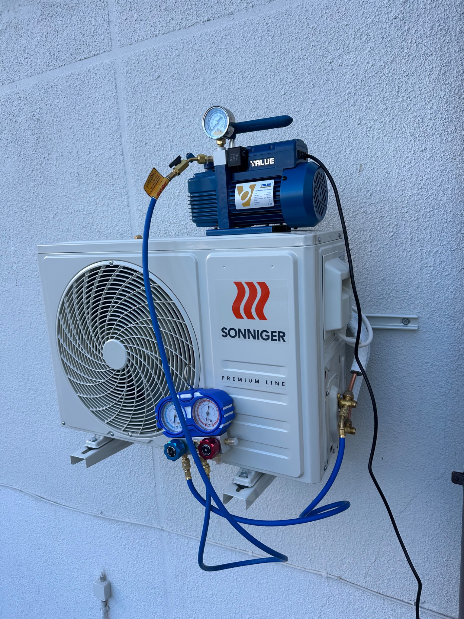 Biała jednostka zewnętrzna klimatyzacji Sonniger Premium Line, podłączona do niebieskiej pompy próżniowej VALUE z manometrem, zamontowana na białej, teksturowanej ścianie.