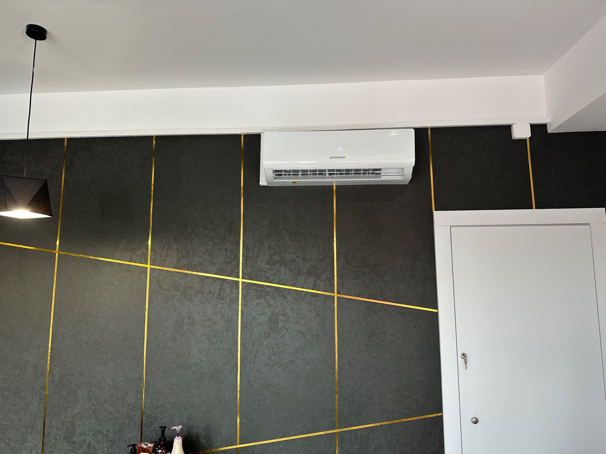 Biały klimatyzator marki Sonniger zamontowany na ciemnej ścianie z geometrycznym wzorem złotych pasków w nowoczesnym wnętrzu.