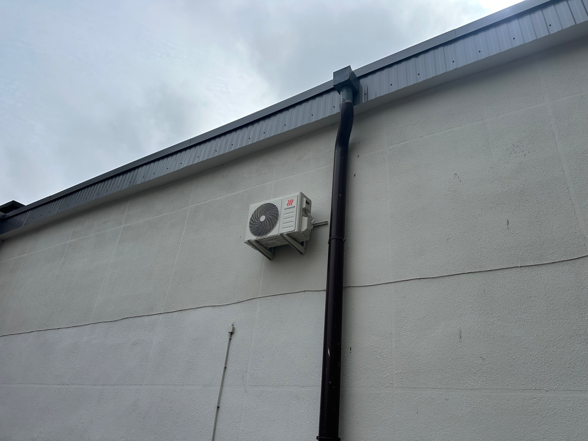 Biała klimatyzacja typu split zamontowana na szarej, teksturowanej ścianie budynku z brązową rynną. Pochmurne niebo w tle.