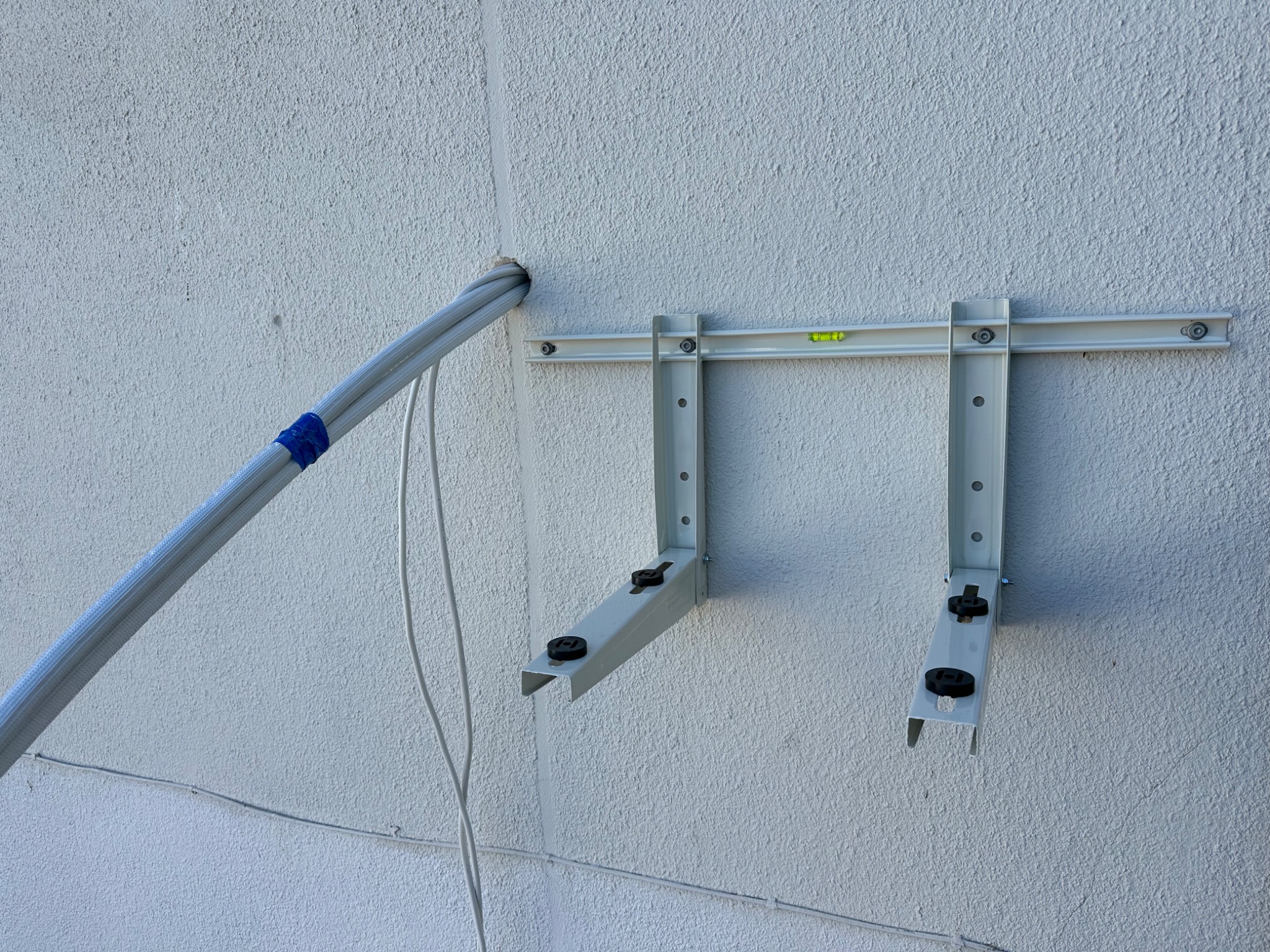 Montaż wspornika klimatyzacji na teksturowanej ścianie. Widoczne przewody w peszlu i poziomica na listwie montażowej. Etap instalacji, przygotowanie do montażu jednostki zewnętrznej.