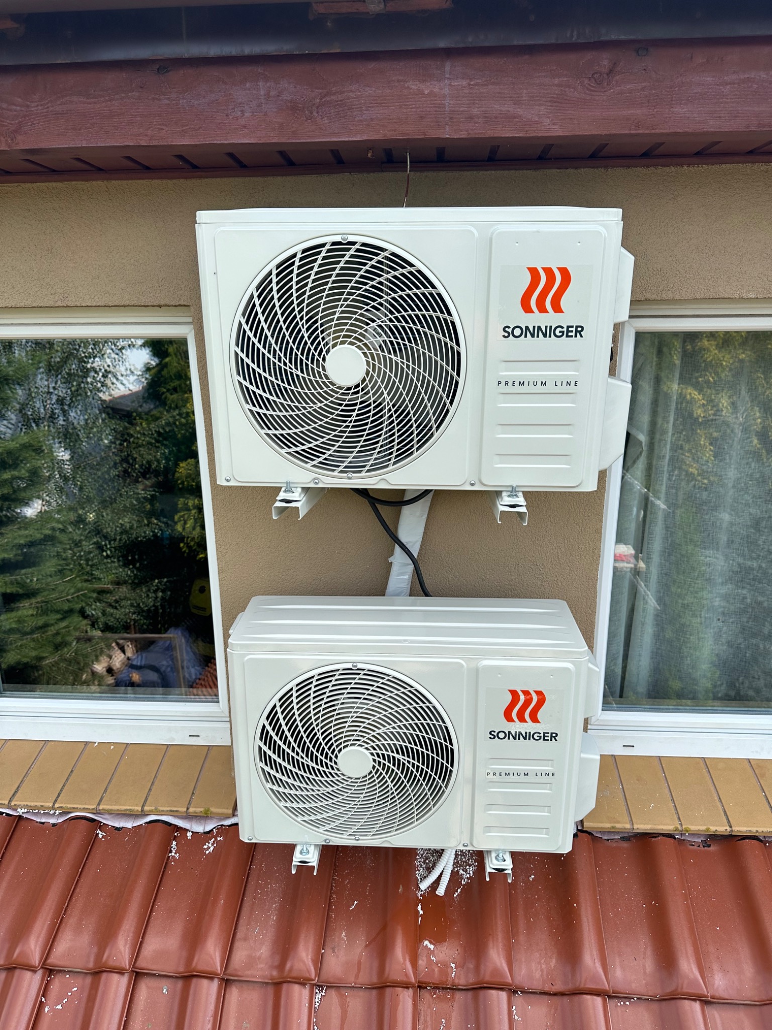 Dwa klimatyzatory marki Sonniger Premium Line zamontowane na elewacji budynku, widoczne okna z odbijającym się drzewem i czerwona dachówka.
