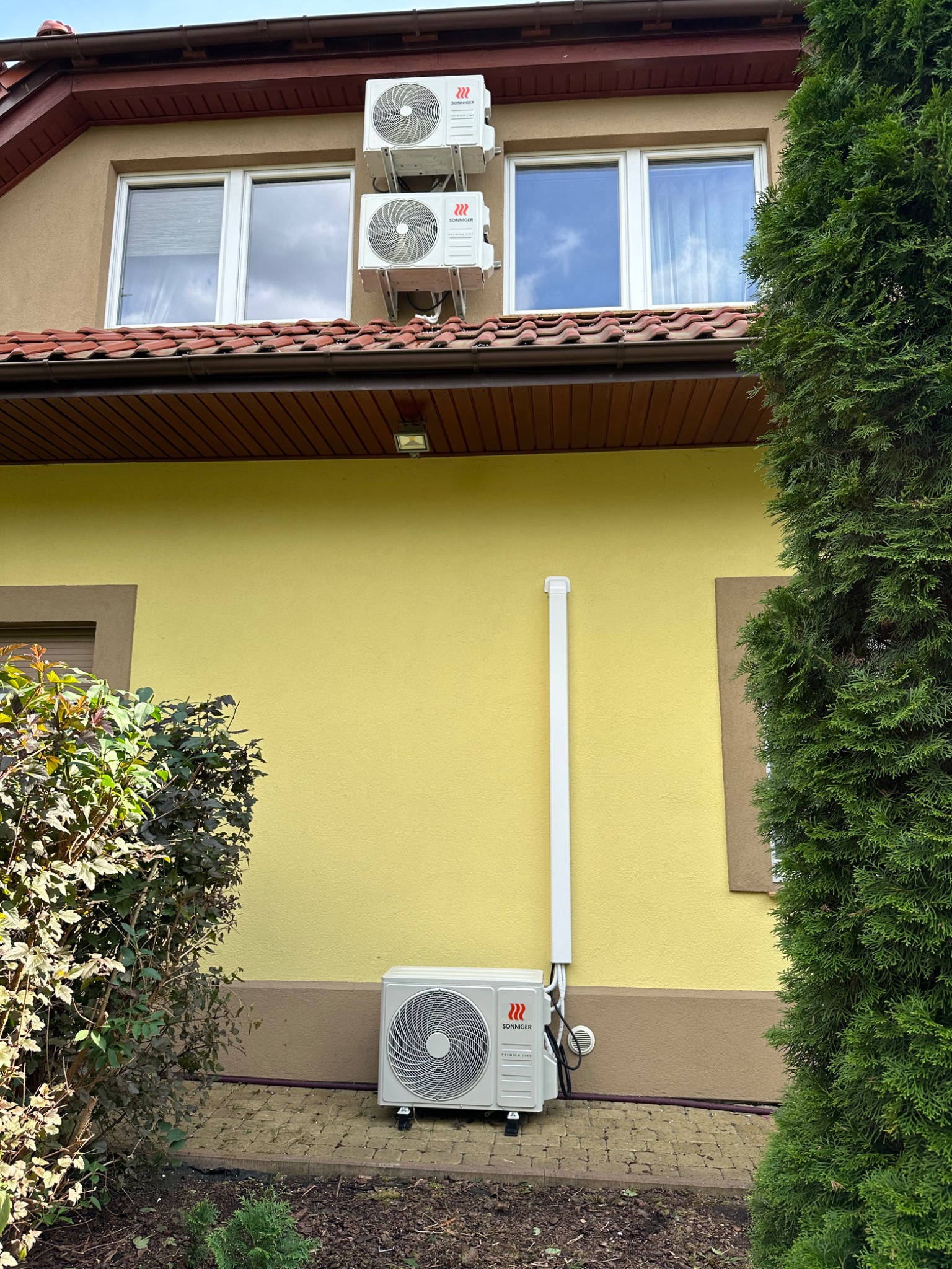 Żółta elewacja domu z trzema klimatyzatorami marki Sonniger: dwa na górze pod oknami i jeden na dole, przy krzewie. Widoczna instalacja odprowadzająca skropliny.