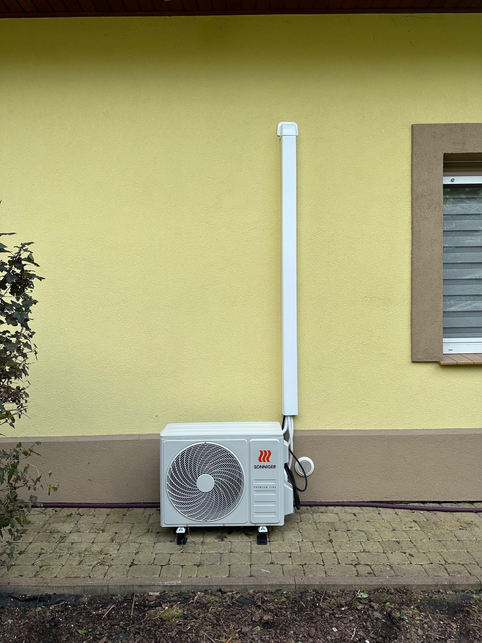 Zewnętrzna jednostka klimatyzacji Sonniger Premium Line, zamontowana na kostce brukowej przy żółtej elewacji budynku z brązowym cokołem i oknem w brązowej ramie.