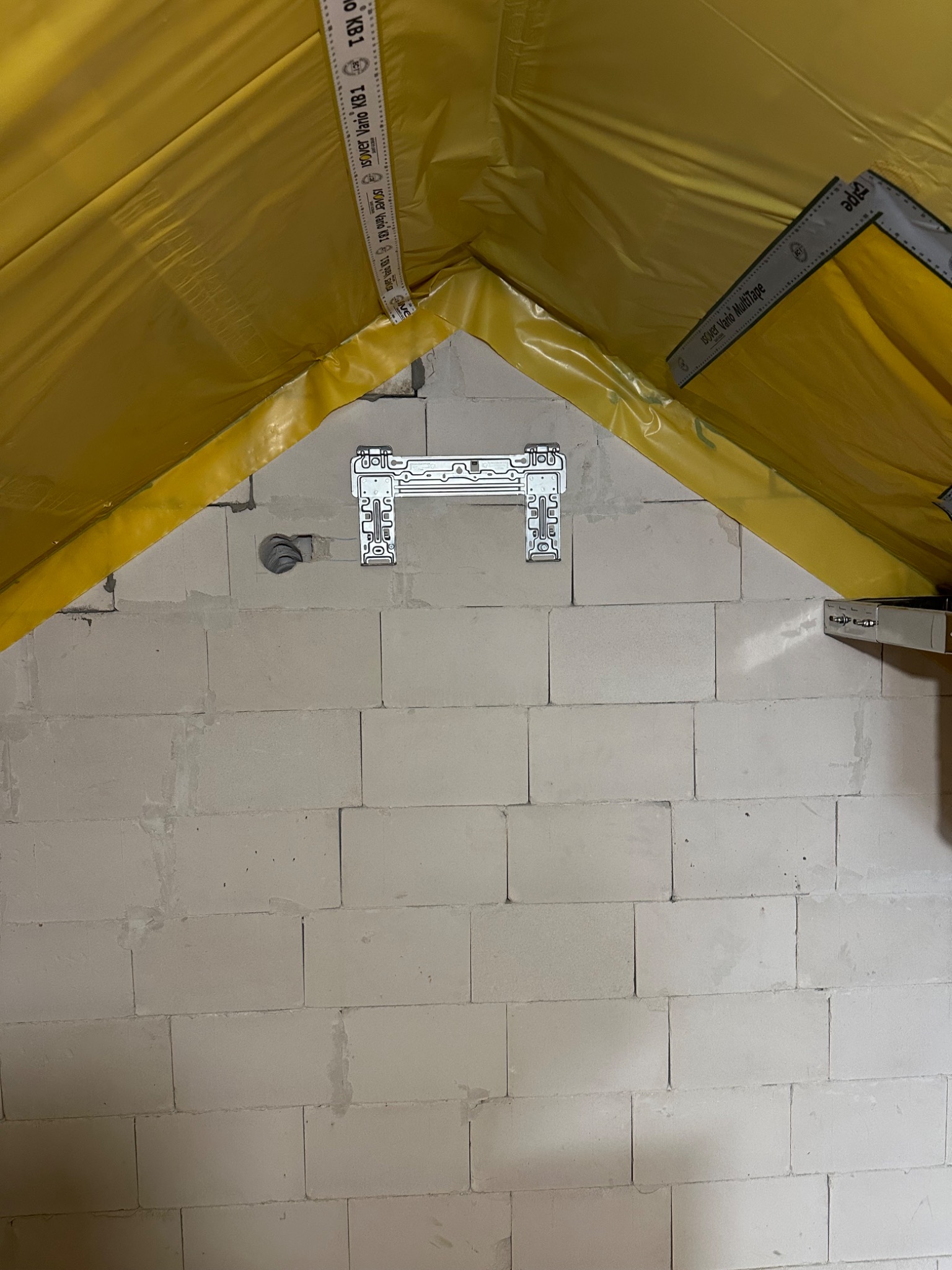 Montaż wspornika klimatyzacji na ścianie z bloczków, widoczna izolacja poddasza i otwór na przewody. Przygotowanie do instalacji klimatyzacji.