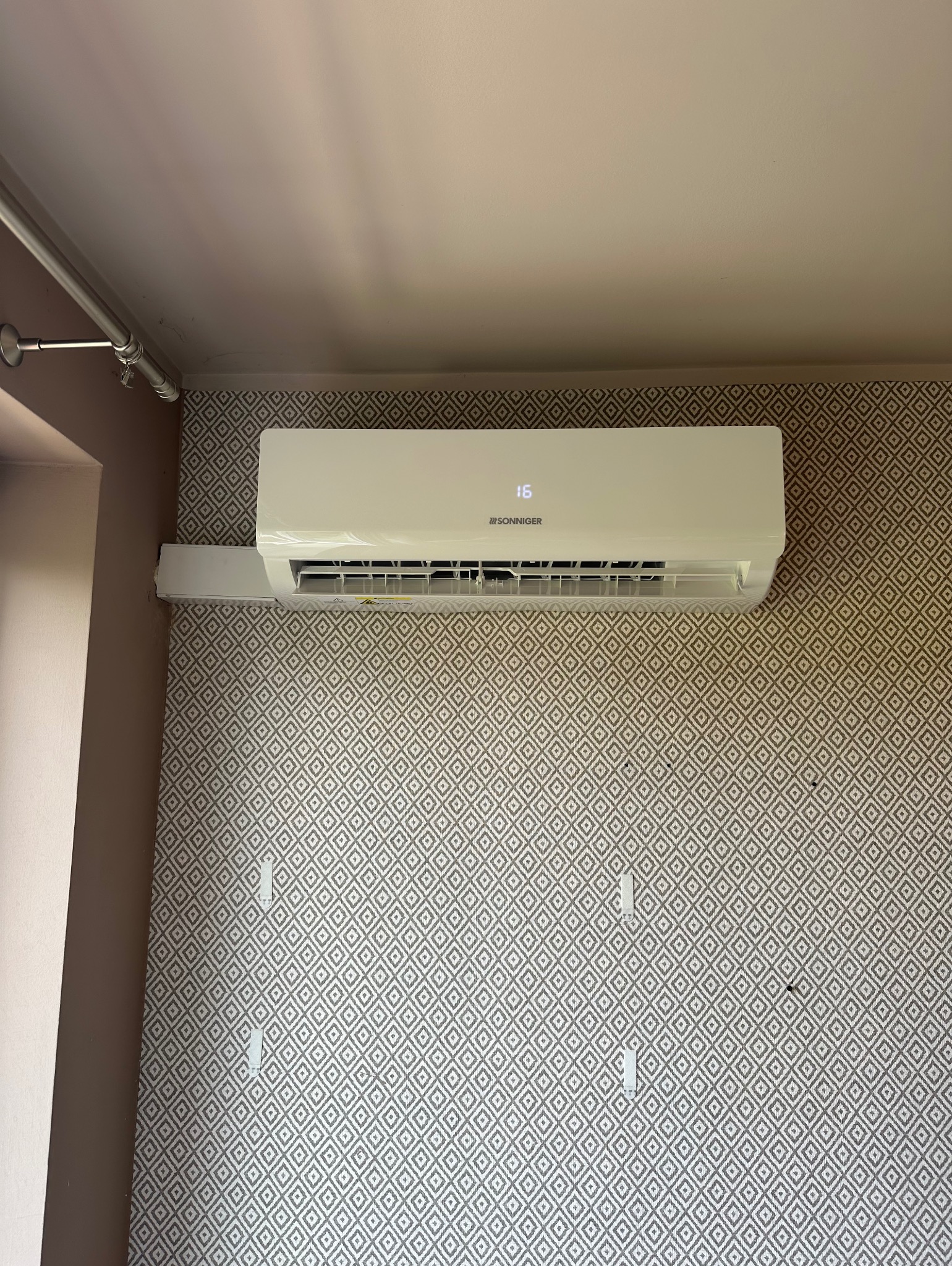 Biała klimatyzacja marki Sonniger zamontowana na wzorzystej ścianie w pomieszczeniu. Widoczne przewody na suficie i fragment ściany w innym kolorze.