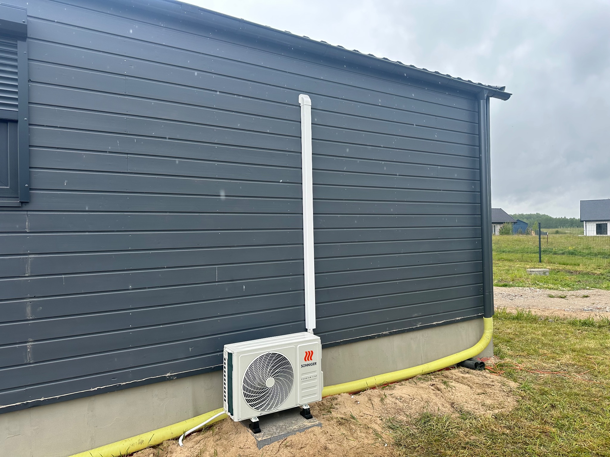 Biały klimatyzator zewnętrzny marki Sonniger zamontowany na szarej elewacji domu z drewnianą okładziną, z widocznymi rurami odprowadzającymi.