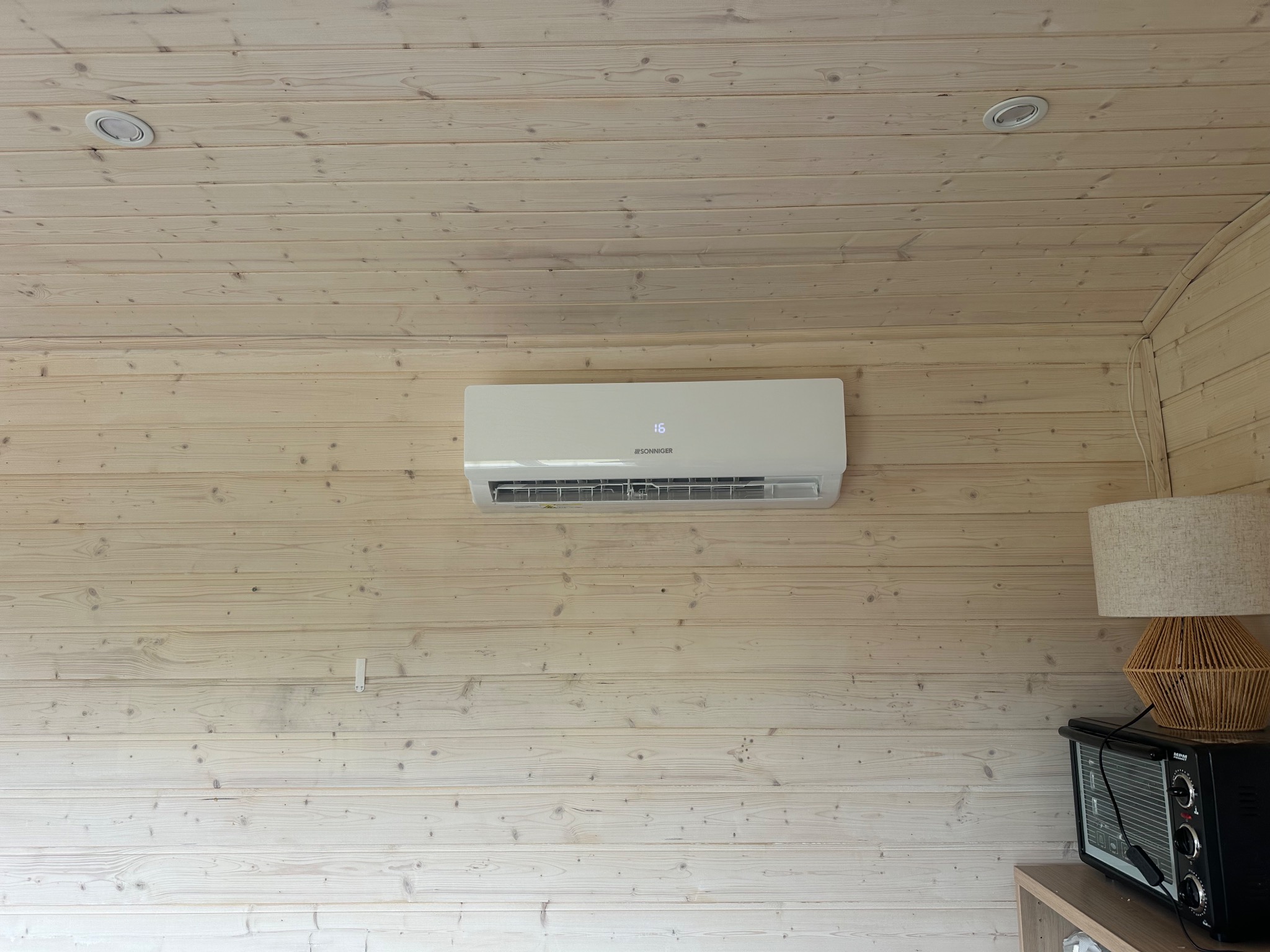 Biała klimatyzacja Sonniger na drewnianej ścianie w jasnym pomieszczeniu z lampą i piekarnikiem na szafce. Widoczne oświetlenie sufitowe.