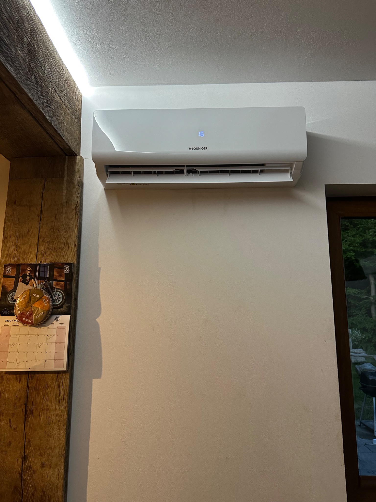 Biały klimatyzator marki Sonniger zamontowany na ścianie w pomieszczeniu, obok drewnianej belki i kalendarza. Widoczny fragment okna.
