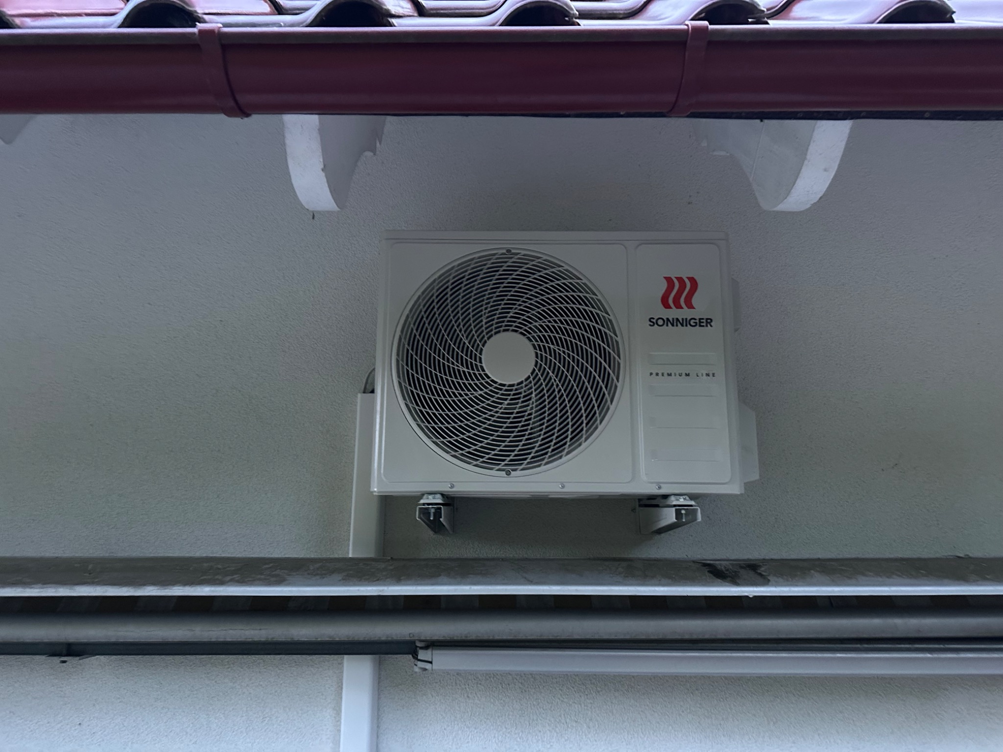 Biała jednostka zewnętrzna klimatyzacji marki Sonniger zamontowana na ścianie budynku z widocznym dachem i rynną. Widok z dołu na zamontowany klimatyzator.