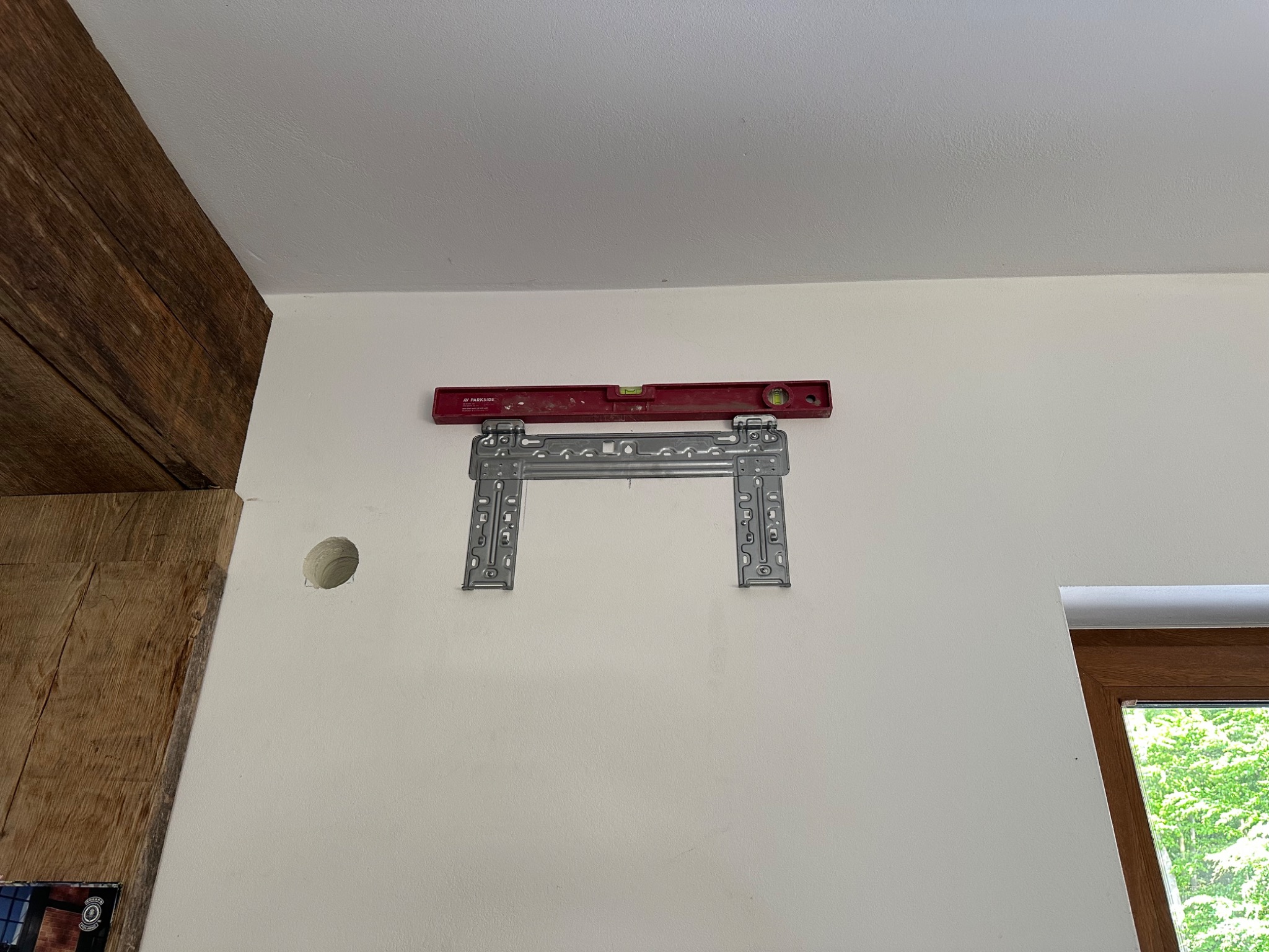 Montaż wspornika pod klimatyzator z poziomicą na ścianie w Gdańsku. Widoczny otwór na instalację. Fragment drewnianej belki stropowej. Okno z zielenią w tle.