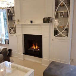 ART-DREW USŁUGI STOLARSKIE NIP 777 177 04 26 - Biała zabudowa kominkowa z przeszklonymi witrynami po bokach, płomienie w czarnym wkładzie kominkowym. Klasyczny salon z elementami dekoracyjnymi.