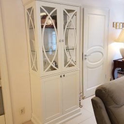 ART-DREW USŁUGI STOLARSKIE NIP 777 177 04 26 - Elegancki, biały kredens z przeszkloną górną częścią i dekoracyjnymi krzyżulcami, stojący w jasnym wnętrzu obok białych drzwi.