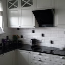 ART-DREW USŁUGI STOLARSKIE NIP 777 177 04 26 - Jasna kuchnia z białymi szafkami, czarnym blatem i okapem. Górne szafki z przeszkleniami w kształcie X. Ściana wyłożona białymi płytkami z czarnymi akcentami.