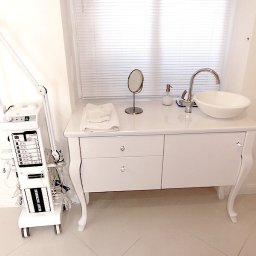 ART-DREW USŁUGI STOLARSKIE NIP 777 177 04 26 - Biała szafka łazienkowa z umywalką nablatową i lustrem, obok biały sprzęt kosmetyczny na kółkach. Ujęcie w jasnym wnętrzu, blisko okna z żaluzjami.