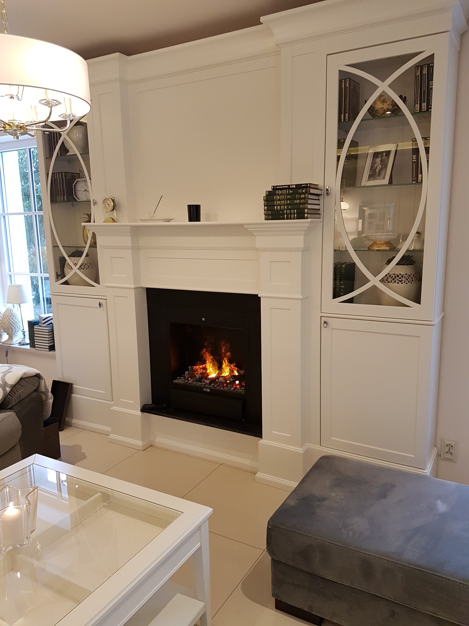 Biała zabudowa kominkowa z przeszklonymi witrynami po bokach, płomienie w czarnym wkładzie kominkowym. Klasyczny salon z elementami dekoracyjnymi.