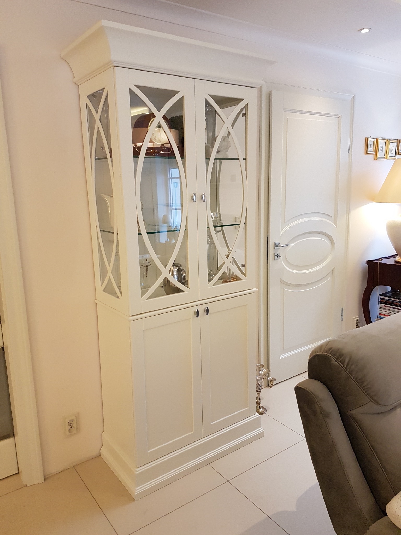 Elegancki, biały kredens z przeszkloną górną częścią i dekoracyjnymi krzyżulcami, stojący w jasnym wnętrzu obok białych drzwi.