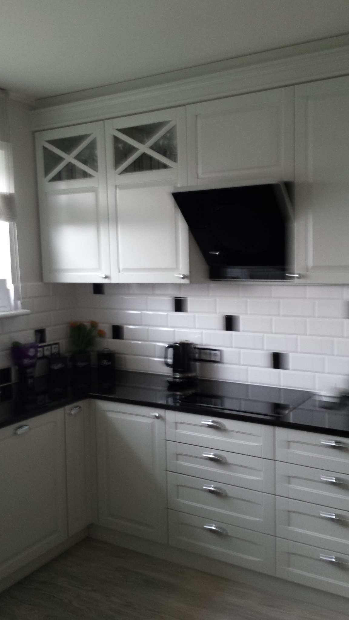 Jasna kuchnia z białymi szafkami, czarnym blatem i okapem. Górne szafki z przeszkleniami w kształcie X. Ściana wyłożona białymi płytkami z czarnymi akcentami.