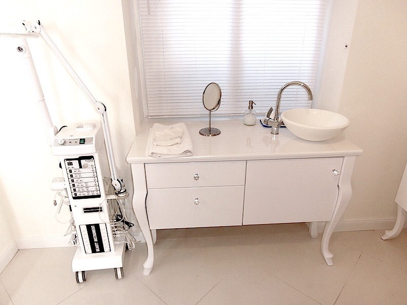 Biała szafka łazienkowa z umywalką nablatową i lustrem, obok biały sprzęt kosmetyczny na kółkach. Ujęcie w jasnym wnętrzu, blisko okna z żaluzjami.