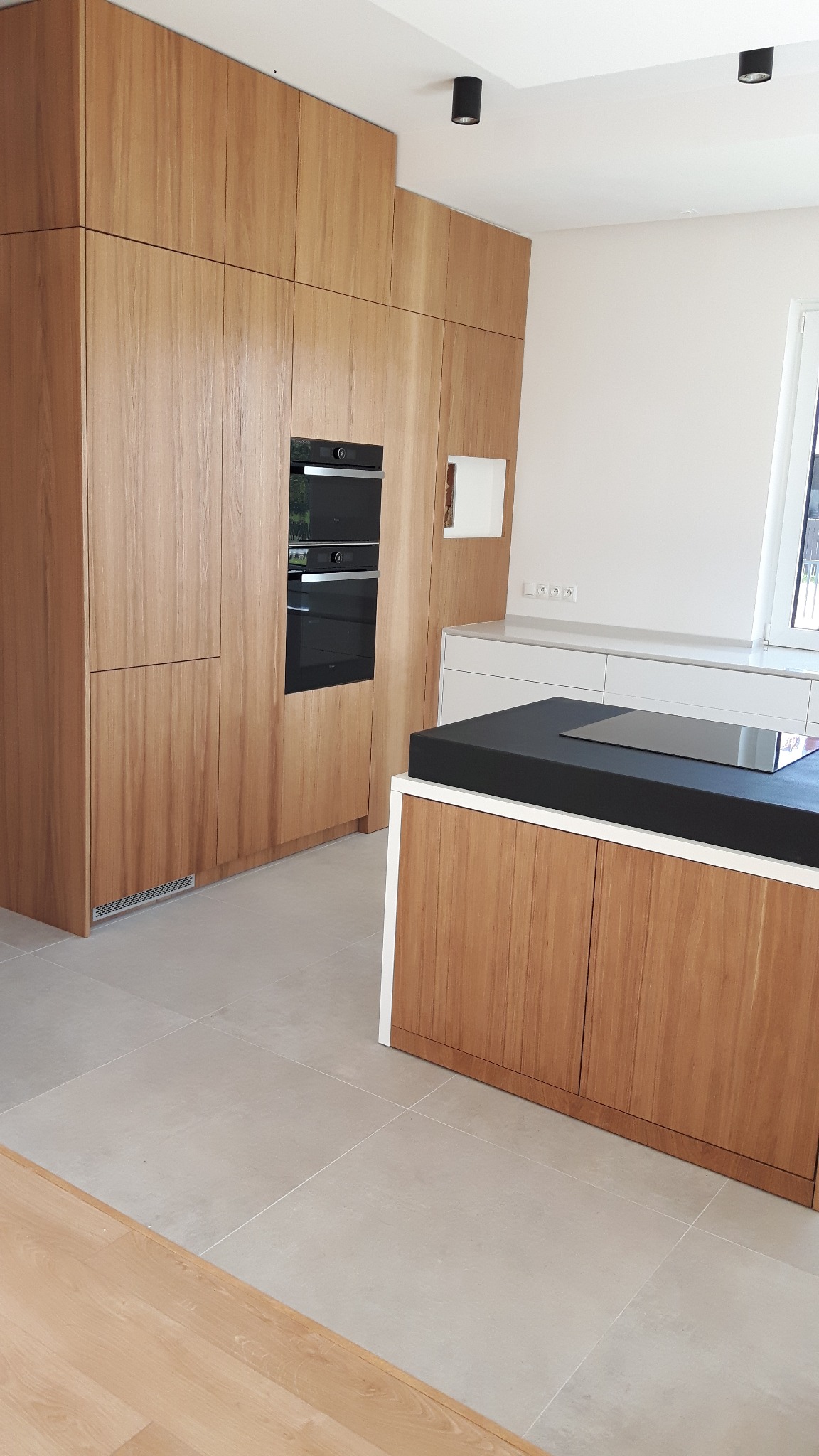 Kuchnia z frontami z jasnego drewna, zabudowana od podłogi do sufitu z wbudowanym piekarnikiem i wyspą kuchenną z czarnym blatem. Minimalistyczne oświetlenie.
