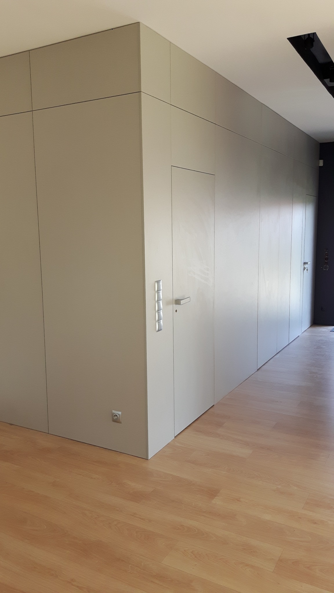 Minimalistyczna zabudowa ściany z ukrytymi drzwiami, wykończona w jasnym odcieniu szarości. Widoczny fragment drewnianej podłogi i gniazdko elektryczne przy ścianie.