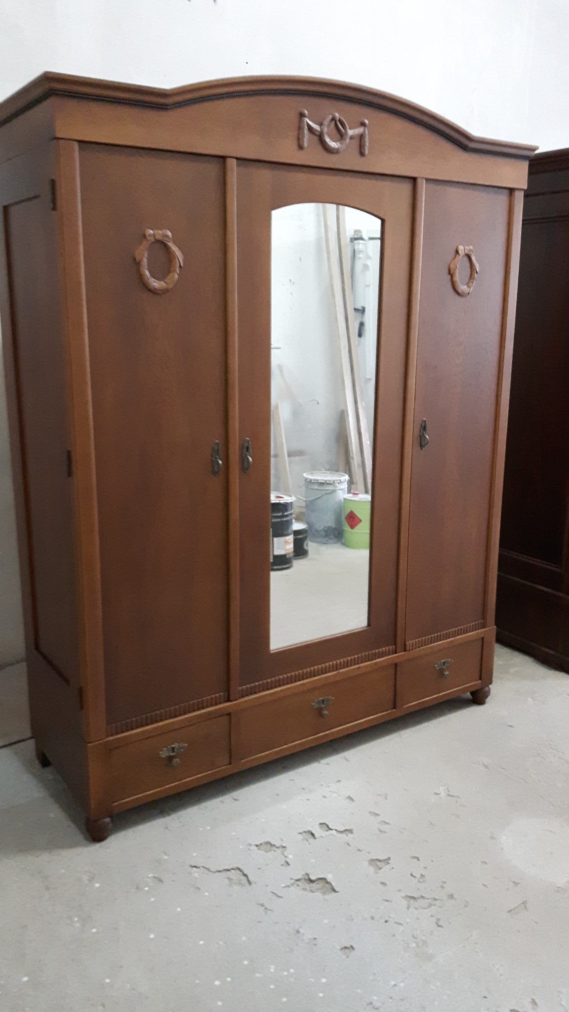 Trzydrzwiowa, używana szafa z lustrem pośrodku, w stylu retro. Na drzwiach dekoracyjne okrągłe uchwyty. Widoczne ślady użytkowania na podłodze.