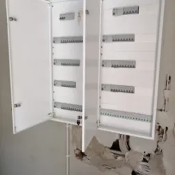 Otwarta, biała skrzynka z bezpiecznikami na ścianie w trakcie montażu. Widoczne kable i instalacja elektryczna. Klucze w zamkach.