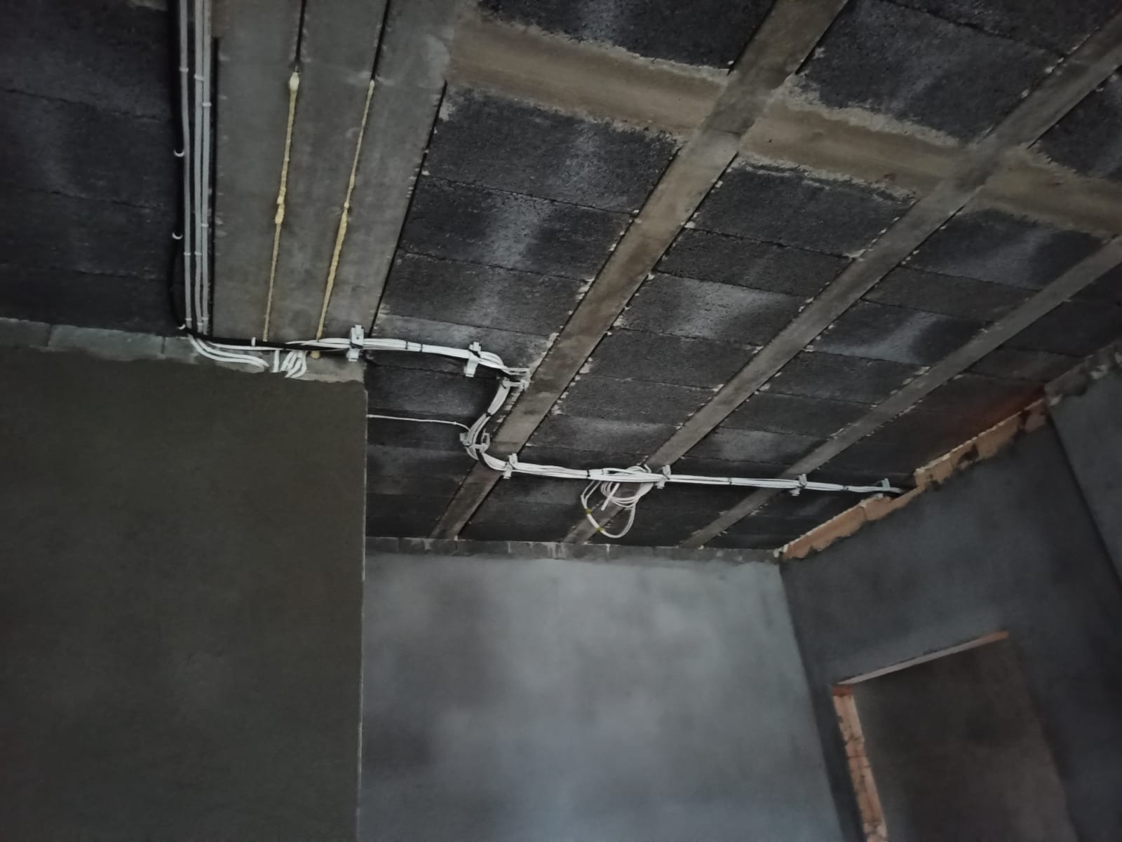 Wnętrze w trakcie remontu z widocznymi przewodami elektrycznymi przymocowanymi do sufitu z betonowych bloczków. Ściany w szarym tynku.