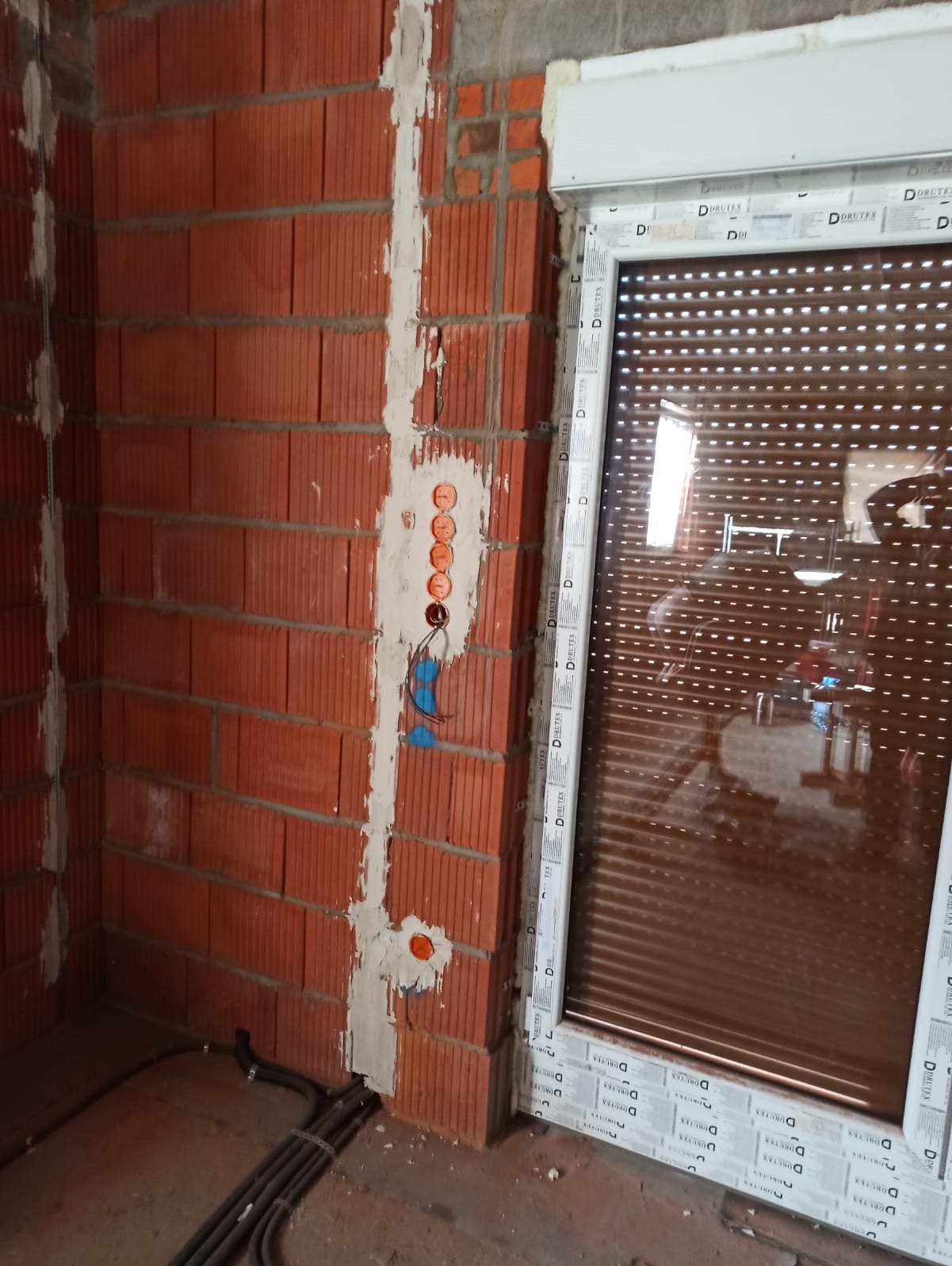 Fragment ściany z czerwonej cegły z widocznymi bruzdami pod instalację elektryczną, pomalowanymi na biało, z puszkami instalacyjnymi i przewodami, obok okna z roletą zewnętrzną.