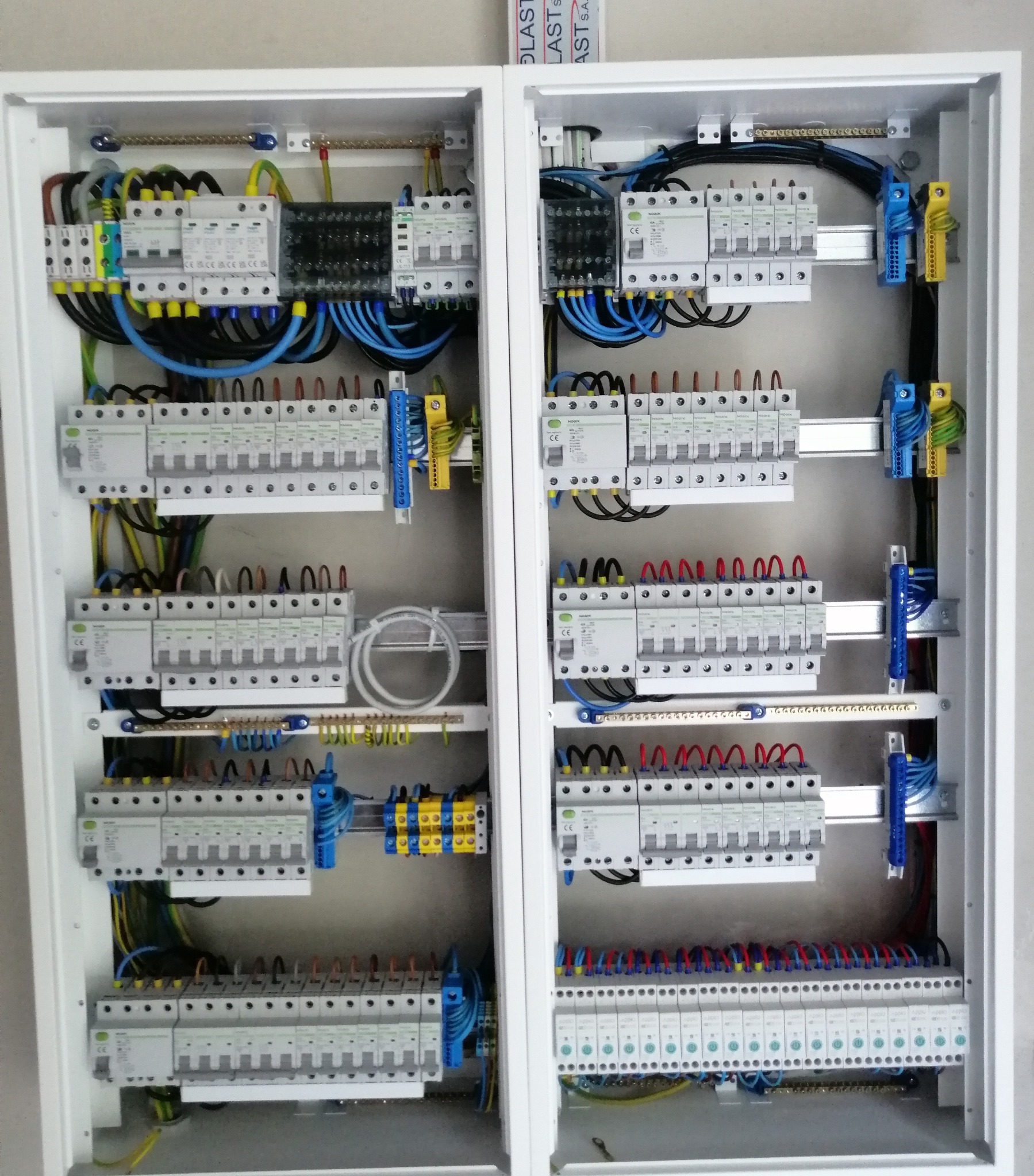 Rozdzielnia elektryczna: dwa panele z bezpiecznikami, okablowaniem i listwami. Widoczne przewody w kolorach niebieskim, żółtym, czarnym i zielonym. Biała obudowa.