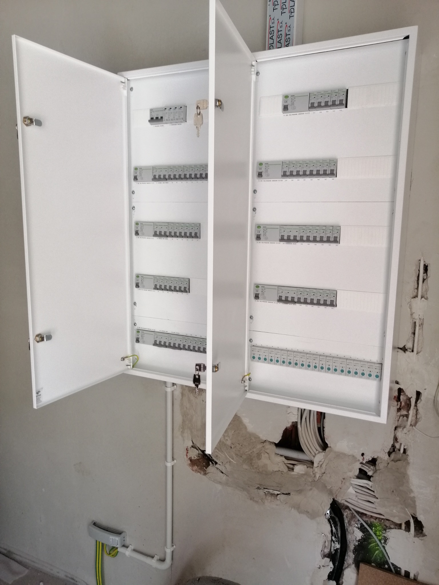 Otwarta, biała skrzynka z bezpiecznikami na ścianie w trakcie montażu. Widoczne kable i instalacja elektryczna. Klucze w zamkach.