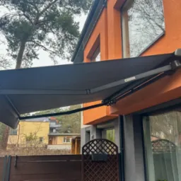 Zewnętrzna, rozkładana markiza w kolorze grafitowym zamontowana nad oknem na pomarańczowej elewacji budynku.