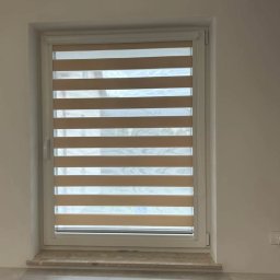 PLISDEKOR Karolina Materka - Zamknięta roleta dzień noc w oknie z białymi ramami, widok z bliska na beżowe pasy rolety i fragment parapetu.