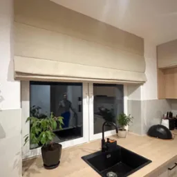 Beżowa roleta rzymska zamontowana w oknie nowoczesnej kuchni z czarnym zlewem i rośliną doniczkową na blacie.