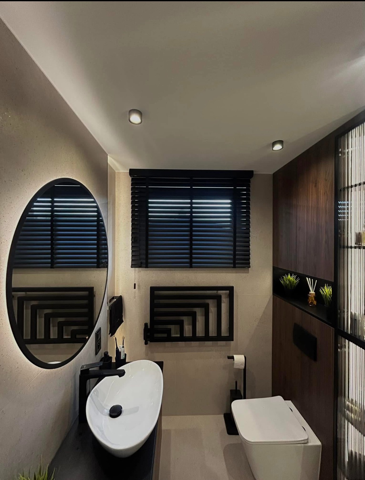 Nowoczesna łazienka w beżach z czarnymi akcentami: poziome żaluzje w oknie, owalne lustro z podświetleniem LED, umywalka nablatowa i grzejnik drabinkowy.