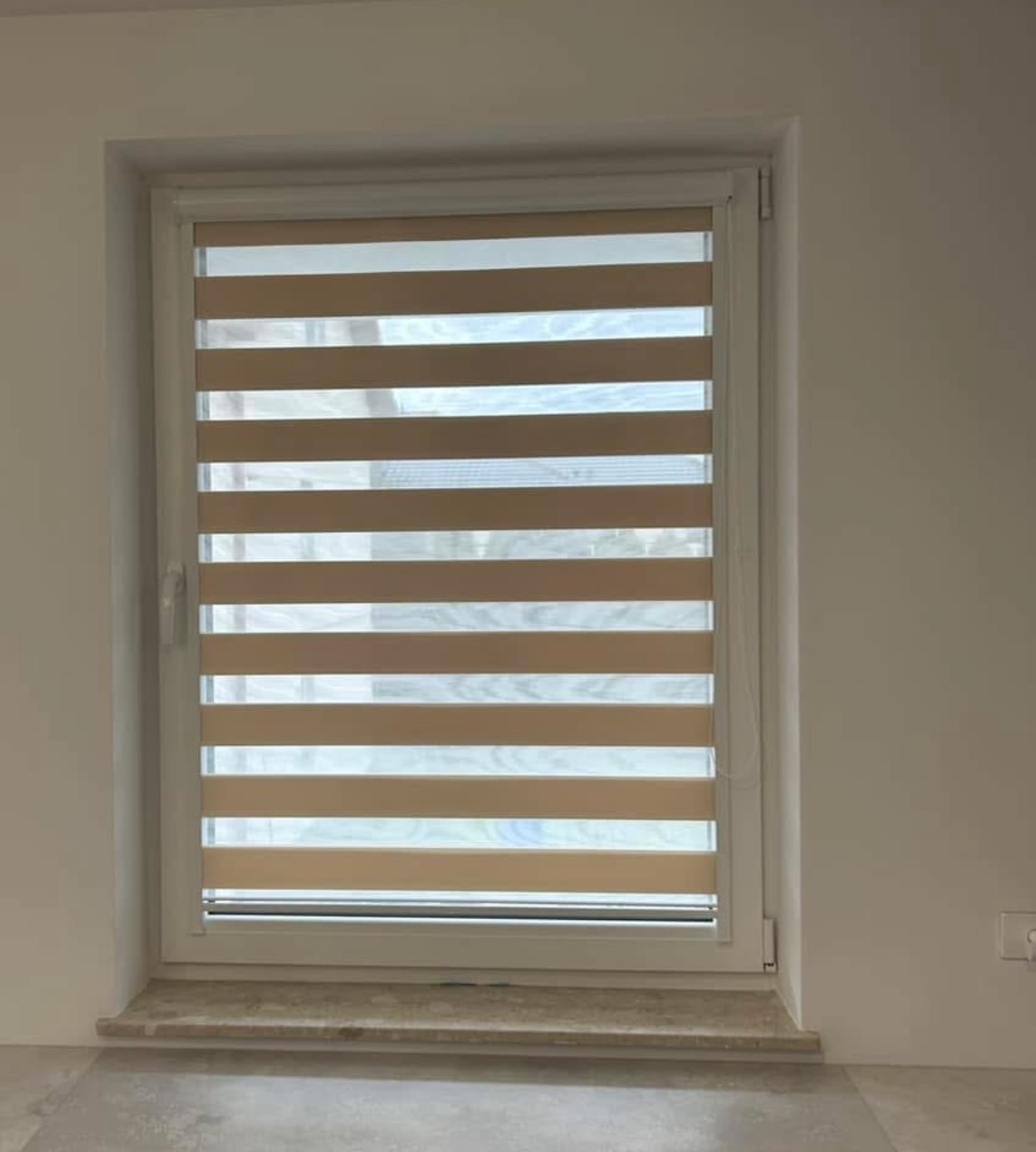 Zamknięta roleta dzień noc w oknie z białymi ramami, widok z bliska na beżowe pasy rolety i fragment parapetu.