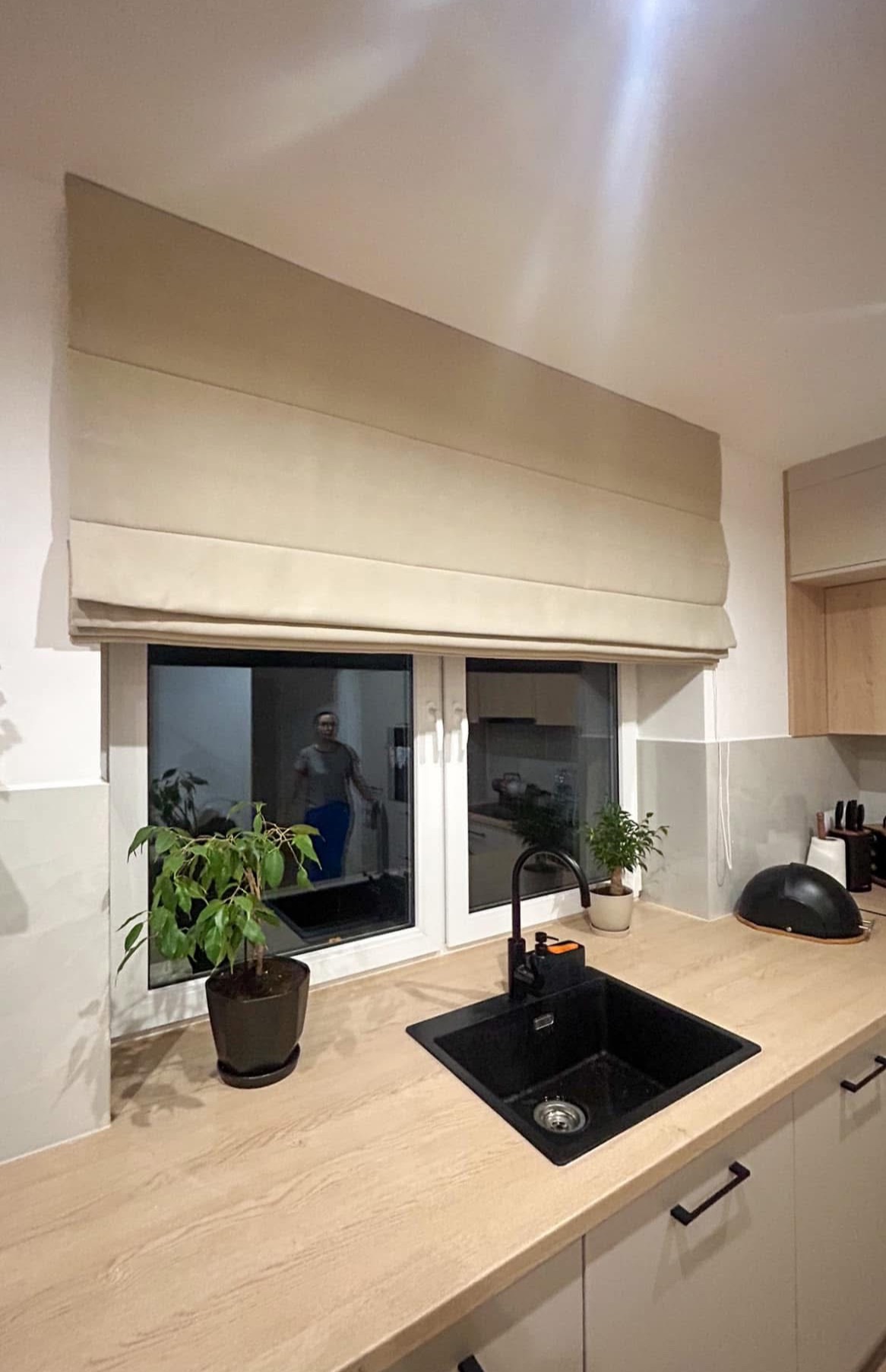 Beżowa roleta rzymska zamontowana w oknie nowoczesnej kuchni z czarnym zlewem i rośliną doniczkową na blacie.