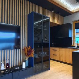 AGNIESZKA WIELGOCKA-PAWLAK NAP studio projektowo-meblarskie - Nowoczesne wnętrze: ściana z drewnianych lameli, niebieska szafka RTV z dekoracjami i szklana witryna, drewniana podłoga, w tle kuchnia z drewnianymi frontami i czarnym blatem.