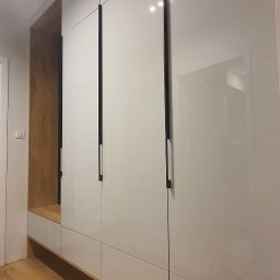 AGNIESZKA WIELGOCKA-PAWLAK NAP studio projektowo-meblarskie - Biała, błyszcząca zabudowa meblowa z ciemnymi, pionowymi uchwytami, wkomponowana we wnękę z drewnianym wykończeniem, widoczna na tle drewnianej podłogi.