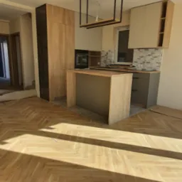 Nowoczesna kuchnia z wyspą w trakcie montażu, widoczne elementy drewniane i szare, parkiet w jodełkę, surowy stan wykończenia pomieszczenia.