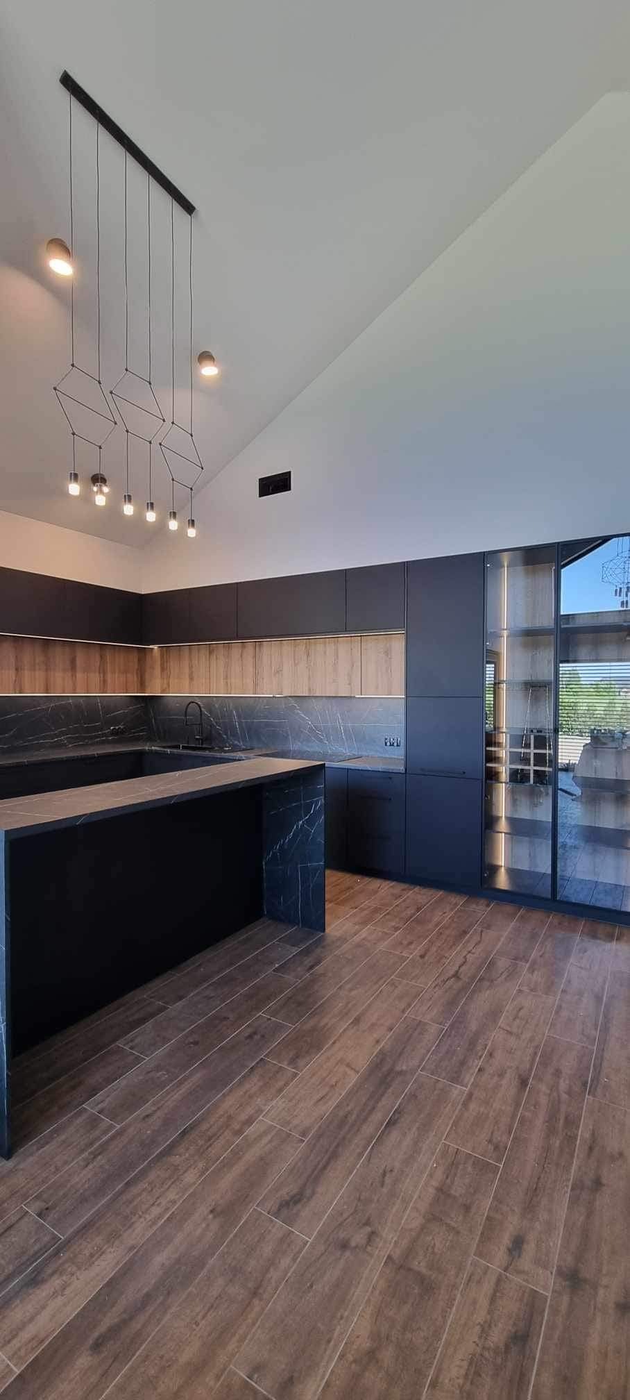 Nowoczesna kuchnia z ciemnymi szafkami, blatem z czarnego marmuru i drewnianą podłogą. Nad wyspą kuchenną zawieszone są geometryczne lampy. Widoczne przeszklone szafki w tle.