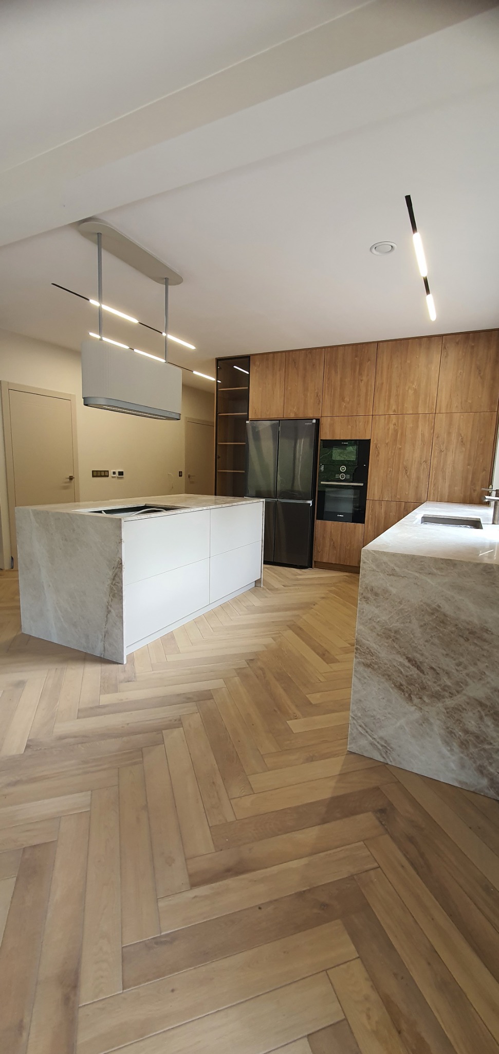 Nowoczesna kuchnia z wyspą i blatem z marmurowym wzorem, drewniane szafki, parkiet w jodełkę i minimalistyczne oświetlenie.