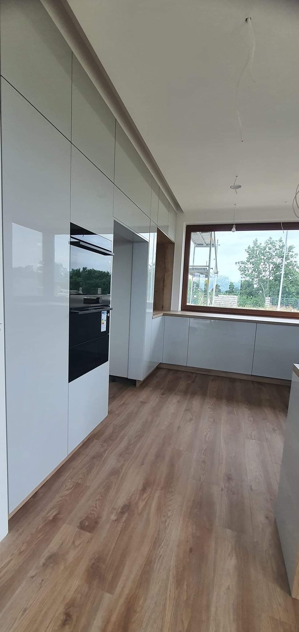 Nowoczesna, jasna kuchnia z białymi, lakierowanymi frontami szafek, drewnianym blatem pod oknem i drewnianą podłogą; wbudowany piekarnik.