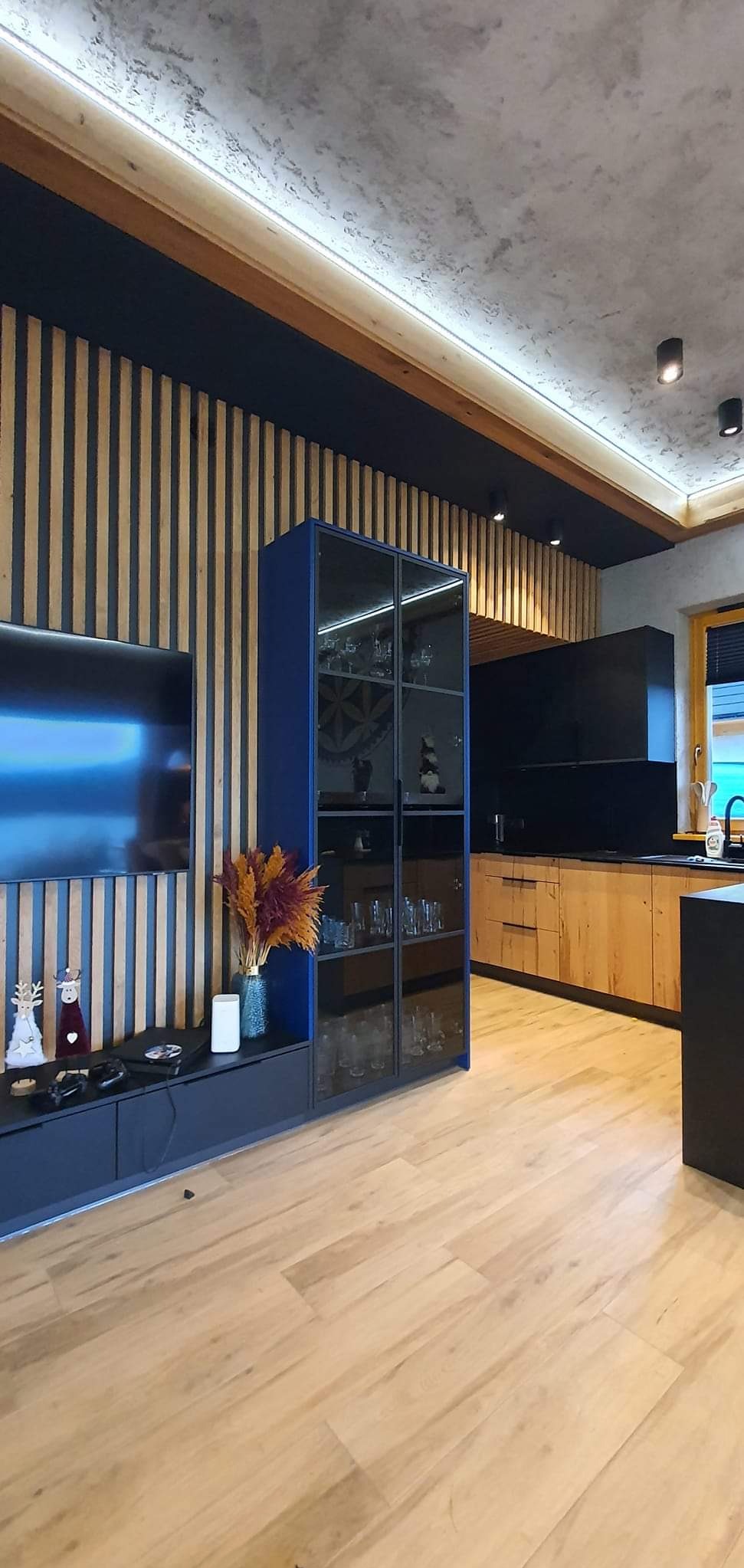 Nowoczesne wnętrze: ściana z drewnianych lameli, niebieska szafka RTV z dekoracjami i szklana witryna, drewniana podłoga, w tle kuchnia z drewnianymi frontami i czarnym blatem.