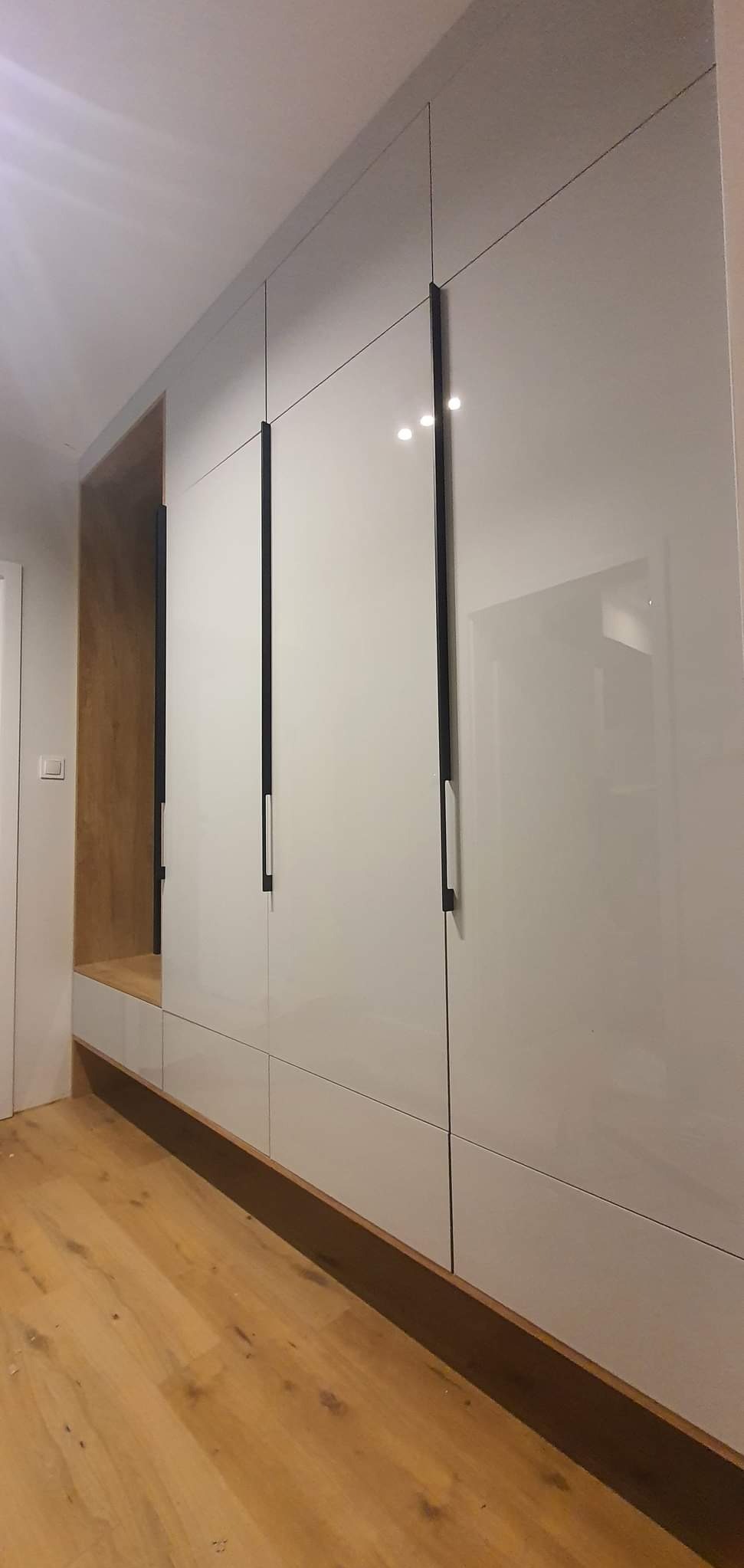 Biała, błyszcząca zabudowa meblowa z ciemnymi, pionowymi uchwytami, wkomponowana we wnękę z drewnianym wykończeniem, widoczna na tle drewnianej podłogi.