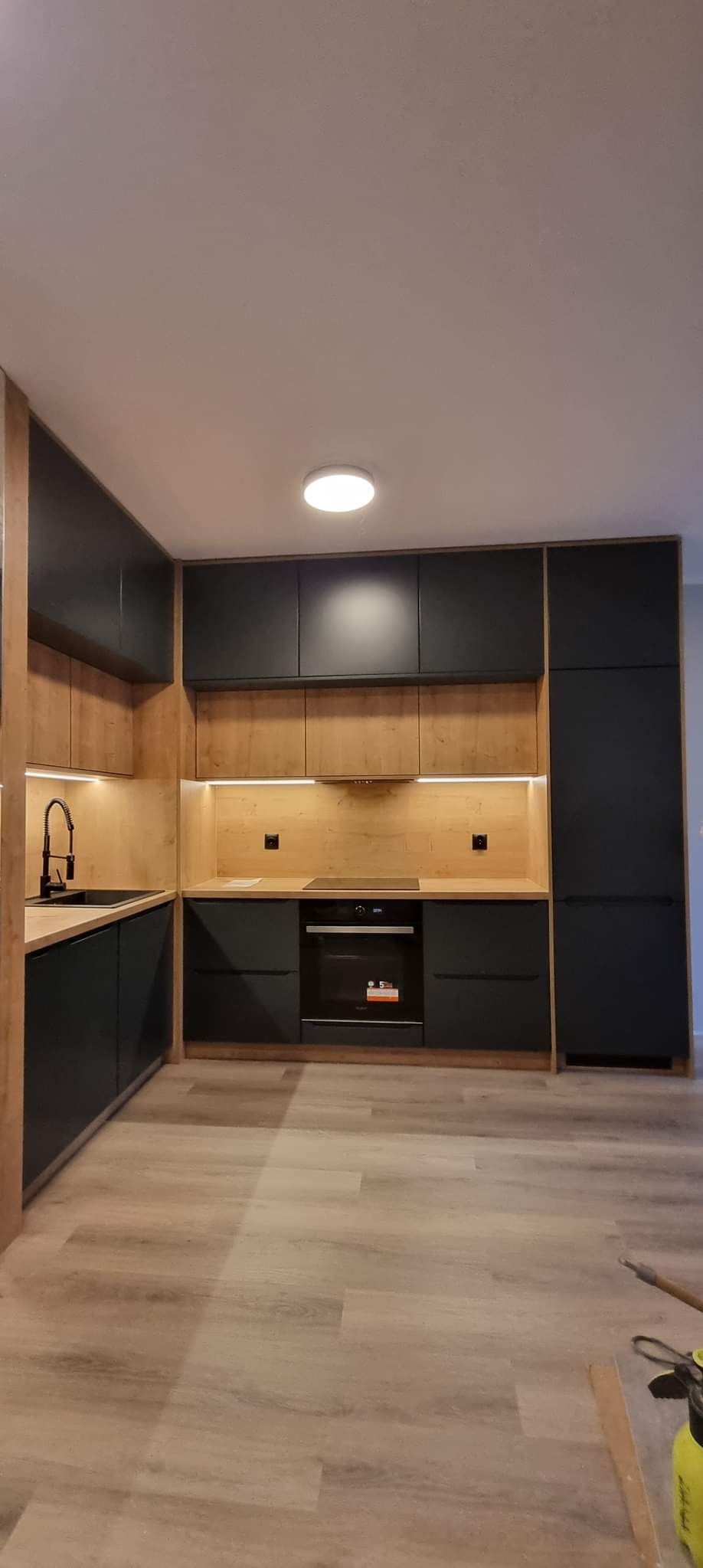 Nowoczesna kuchnia z czarnymi szafkami i drewnianymi elementami, oświetleniem LED pod szafkami wiszącymi i jasną podłogą, widok na fragment podłogi z narzędziami.