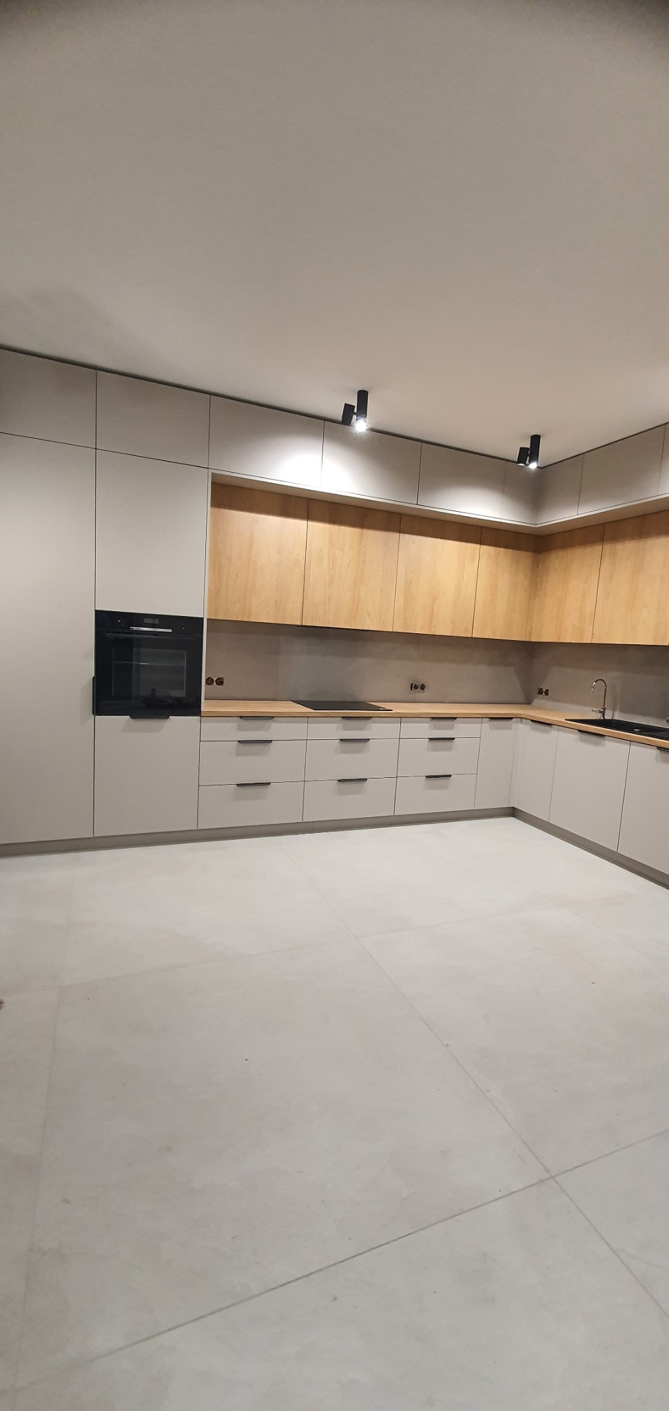 Nowoczesna, jasna kuchnia w Krępice z zabudową w odcieniach szarości i drewna, oświetlona czarnymi reflektorami sufitowymi, z minimalistycznymi uchwytami szuflad i wbudowanym piekarnikiem.