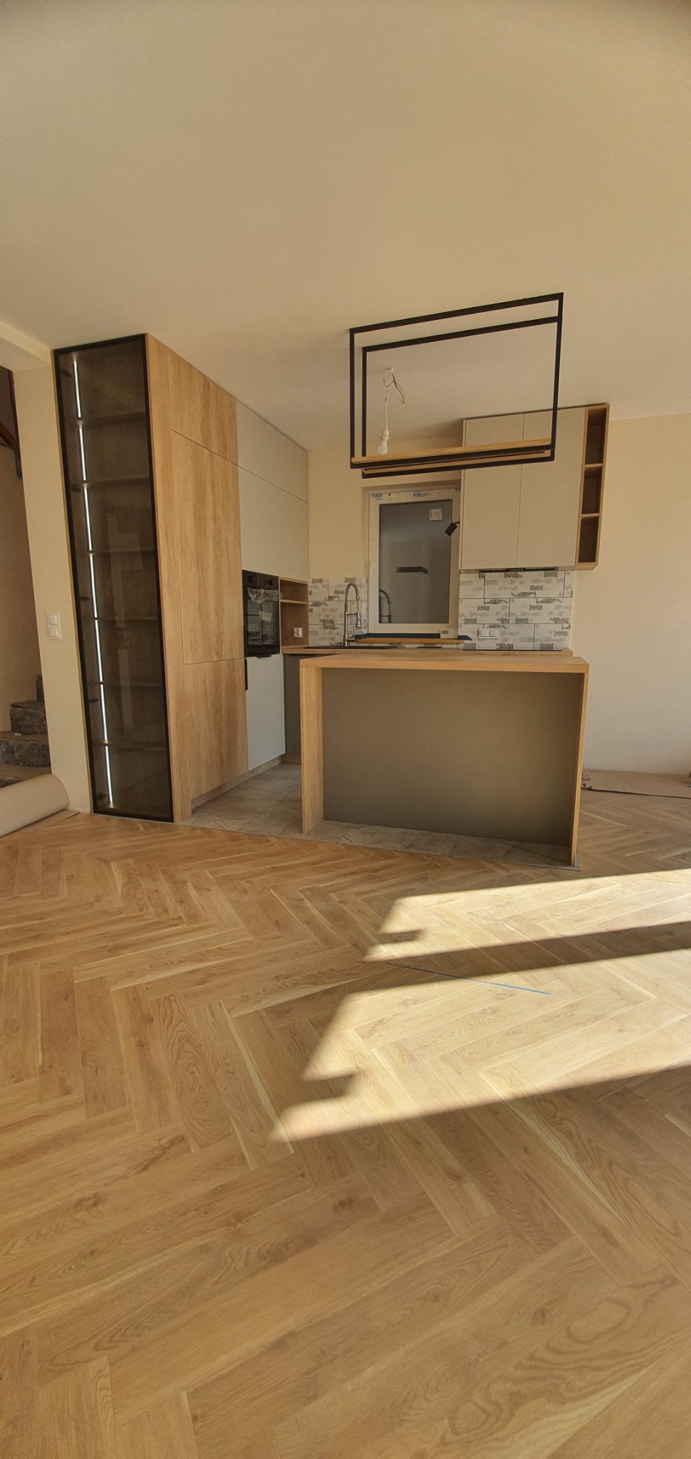 Kuchnia w trakcie montażu, z elementami zabudowy w odcieniach drewna i beżu, wyspą kuchenną i parkietem ułożonym w jodełkę, widoczne niedokończone prace wykończeniowe.