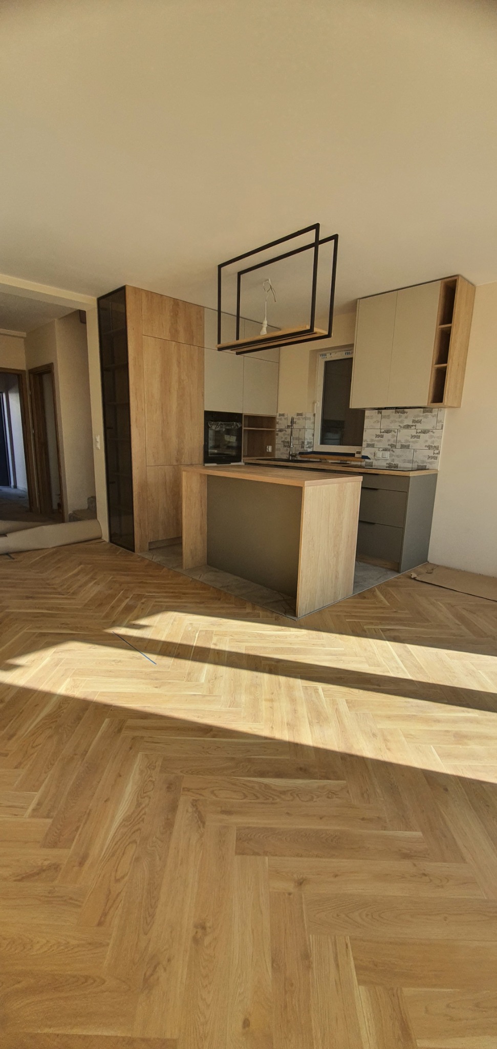 Nowoczesna kuchnia z wyspą w trakcie montażu, widoczne elementy drewniane i szare, parkiet w jodełkę, surowy stan wykończenia pomieszczenia.